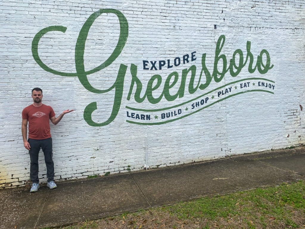 The Author, Wesley Miller, standing with his back facing a wall that has the words: "Explore Greensboro. Learn, Build, Shop, Play, Eat, Enjoy," written on it. 